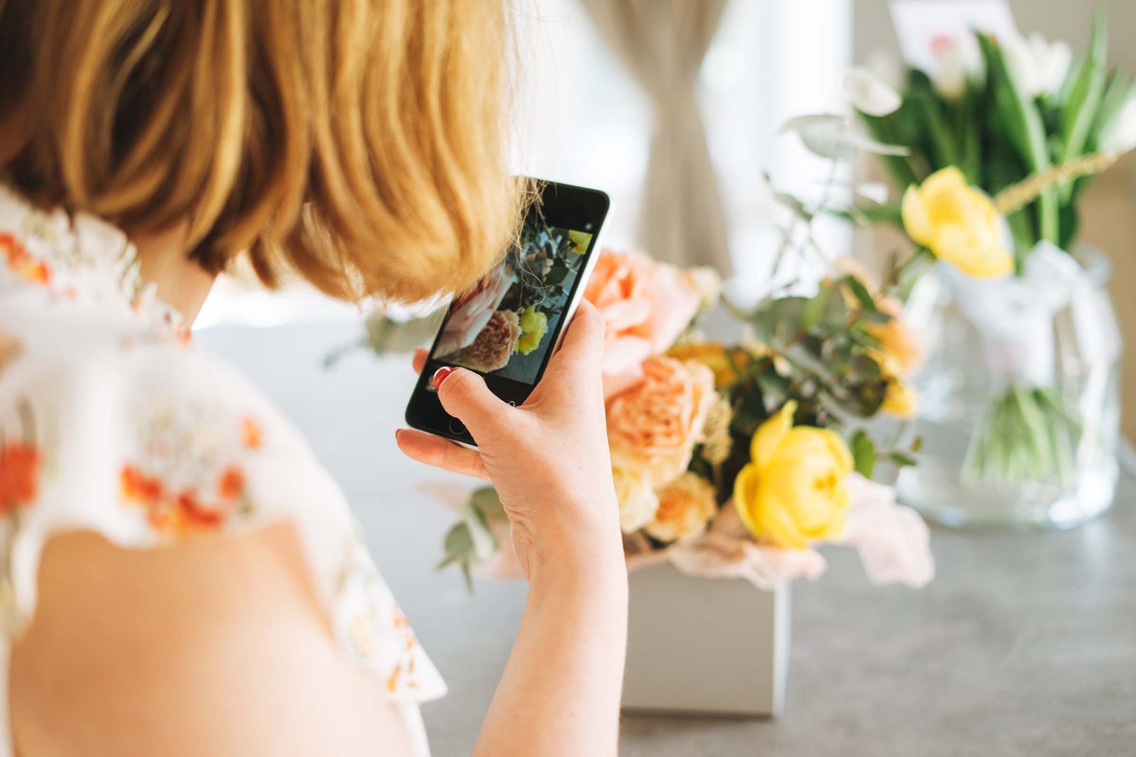 Frau fotografiert mit dem Smartphone eine wei&szlig;e Blumenbox mit Rosen und Tulpen